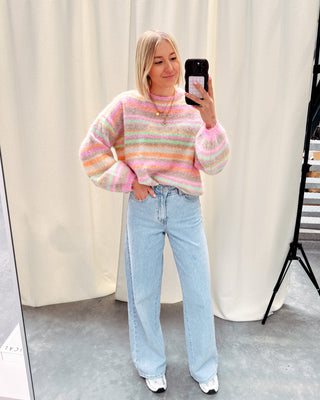 Model draagt een jeansbroek met daarop een regenboog trui in pastelkleurtjes en New Balances.