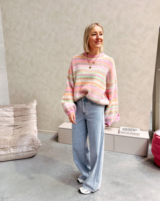 Model draagt een jeansbroek met daarop een regenboog trui in pastelkleurtjes.