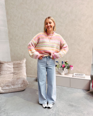 Model draagt een jeansbroek met daarop een regenboog trui in pastelkleurtjes.