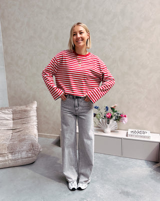 Model draagt een longsleeve met rode en grijze streepjes op een grijze jeansbroek met stretch.