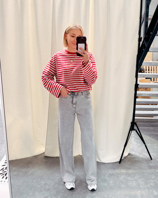 Model draagt een longsleeve met rode en grijze streepjes op een grijze jeansbroek met stretch.
