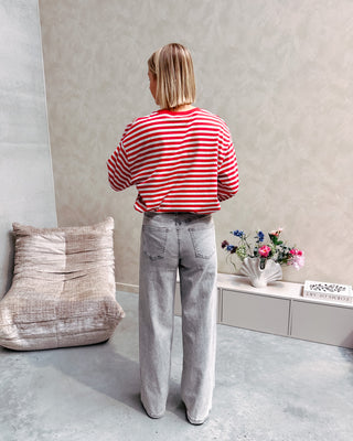 Model draagt een longsleeve met rode en grijze streepjes op een grijze jeansbroek met stretch.