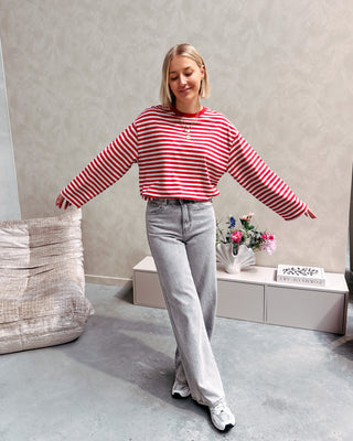 Model draagt een longsleeve met streepjes in rood en grijs op een grijze rechte jeansbroek met veel stretch.