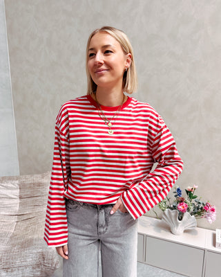 Model draagt een longsleeve met streepjes in rood en grijs op een grijze rechte jeansbroek met veel stretch.