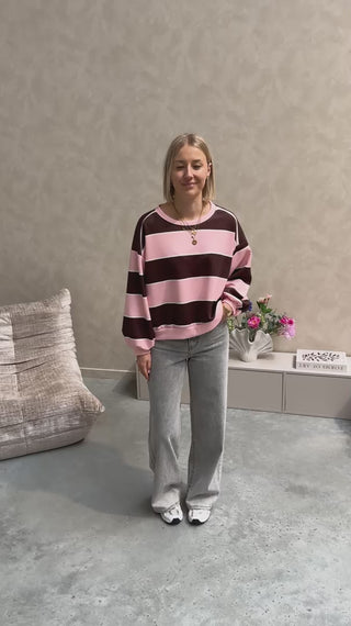 Model draagt een grijze jeansbroek met daarop een gestreepte sweater in de kleuren roos en bruin.
