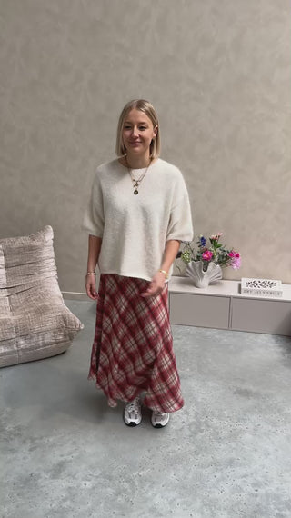 Model draagt een lange rok met ruitjes n oranje en wit, met daarop een witte trui met korte mouwen en achteraan kanten stof.