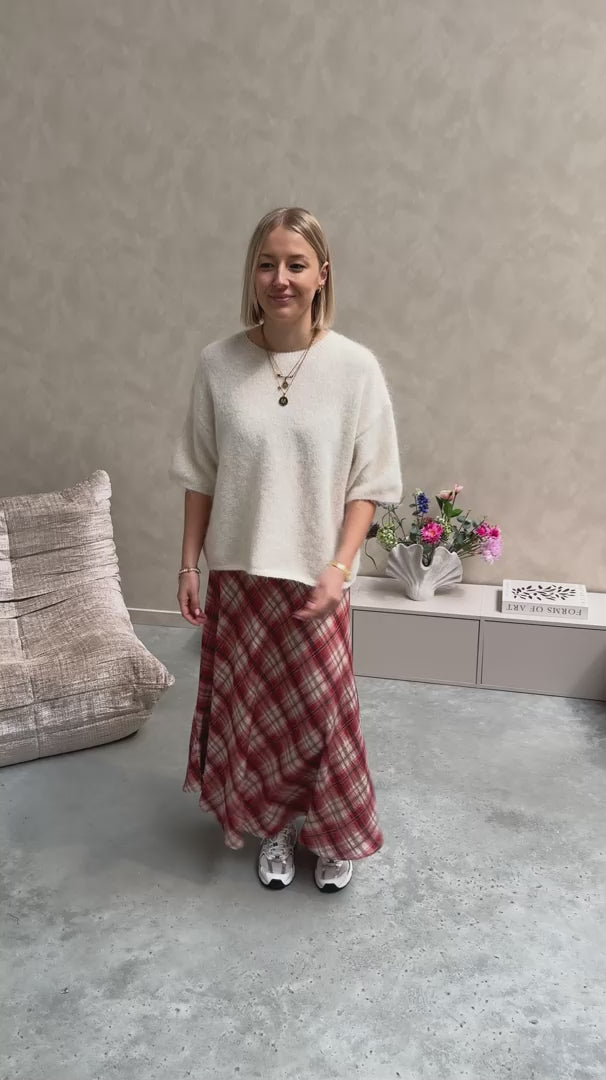 Model draagt een lange rok met ruitjes n oranje en wit, met daarop een witte trui met korte mouwen en achteraan kanten stof.