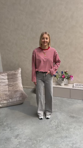 Model draagt een longsleeve met streepjes in rood en grijs op een grijze rechte jeansbroek met veel stretch.