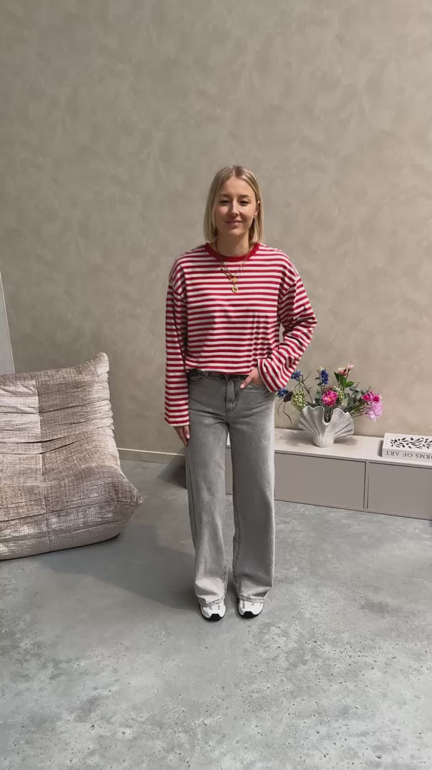 Model draagt een longsleeve met streepjes in rood en grijs op een grijze rechte jeansbroek met veel stretch.
