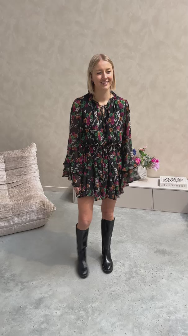 Model draagt een zwarte jurk met een bloemenpatroon. De jurk is getailleerd met een lintje en daaronder draagt ze lange zwarte botten.