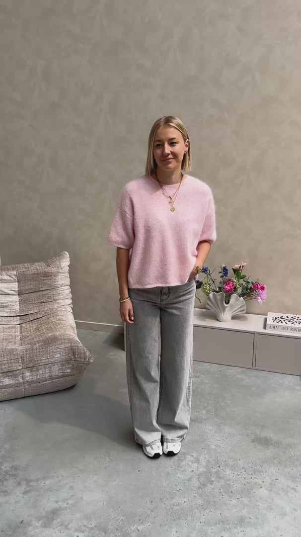 Model draagt een roze trui met korte mouwen en achteraan een kanten stof op een grijze rechte jeansbroek.
