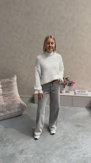Model draagt een grijze jeansbroek met daarop een witte basic coltrui.