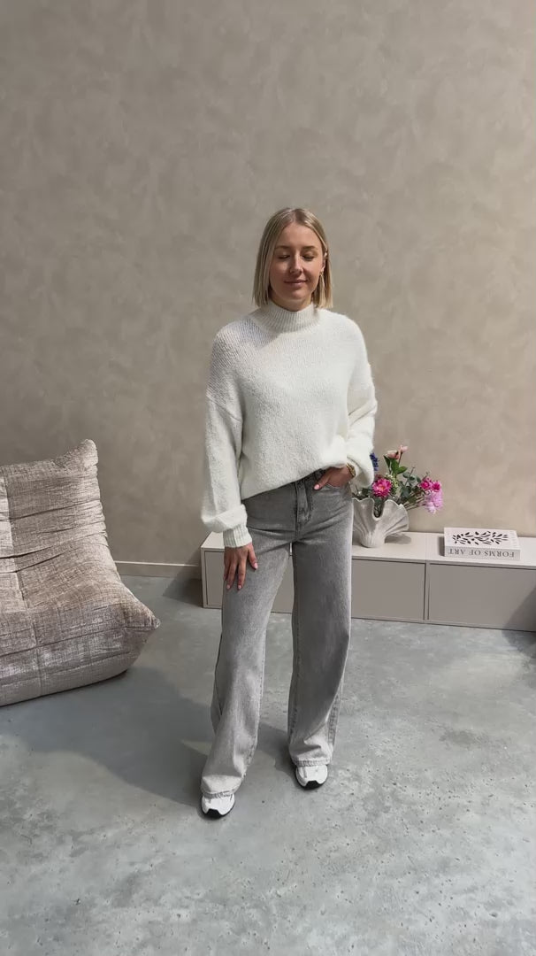Model draagt een grijze jeansbroek met daarop een witte basic coltrui.
