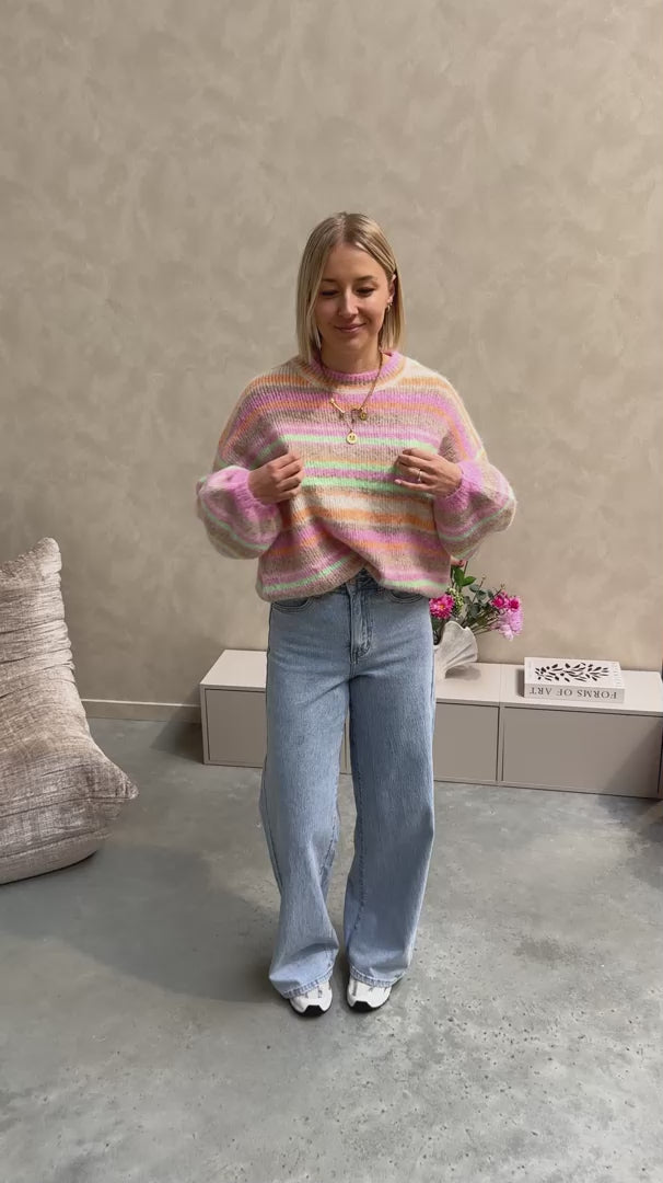Model draagt een jeansbroek met daarop een regenboog trui in pastelkleurtjes.