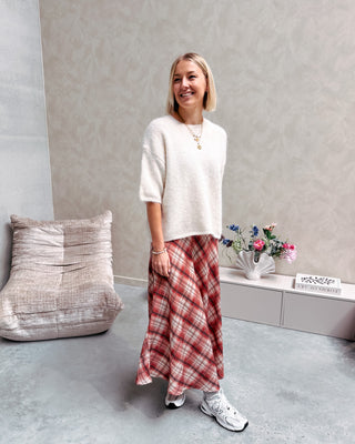 Model draagt een lange rok met ruitjes n oranje en wit, met daarop een witte trui met korte mouwen en achteraan kanten stof.