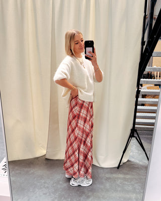 Model draagt een lange rok met ruitjes n oranje en wit, met daarop een witte trui met korte mouwen en achteraan kanten stof.