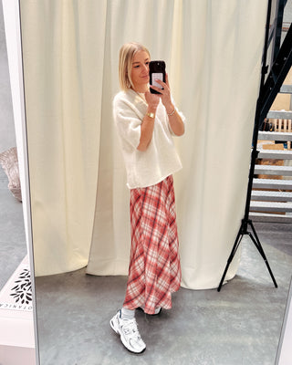 Model draagt een lange rok met ruitjes n oranje en wit, met daarop een witte trui met korte mouwen en achteraan kanten stof.