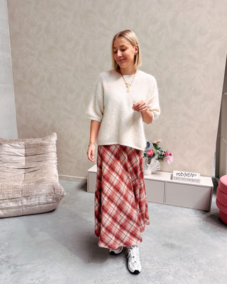 Model draagt een lange rok met ruitjes n oranje en wit, met daarop een witte trui met korte mouwen en achteraan kanten stof.