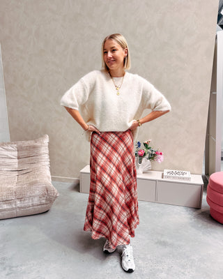 Model draagt een lange rok met ruitjes n oranje en wit, met daarop een witte trui met korte mouwen en achteraan kanten stof.