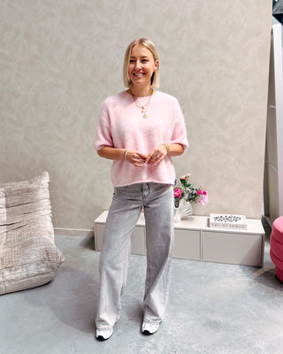 Model draagt een roze trui met korte mouwen en achteraan een kanten stof op een grijze rechte jeansbroek.