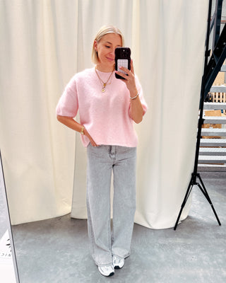 Model draagt een roze trui met korte mouwen en achteraan een kanten stof op een grijze rechte jeansbroek.