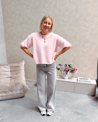 Model draagt een roze trui met korte mouwen en achteraan een kanten stof op een grijze rechte jeansbroek.