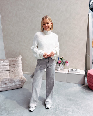 Model draagt een grijze jeansbroek met daarop een witte basic coltrui.
