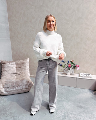 Model draagt een grijze jeansbroek met daarop een witte basic coltrui.