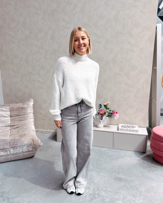 Model draagt een grijze jeansbroek met daarop een witte basic coltrui.