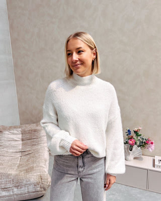 Model draagt een grijze jeansbroek met daarop een witte basic coltrui.