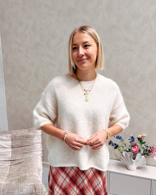 Model draagt een witte trui met korte mouwen en achteraan een kanten stof, op een lange rechte rok met ruitjespatroon.