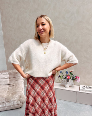 Model draagt een witte trui met korte mouwen en achteraan een kanten stof, op een lange rechte rok met ruitjespatroon.