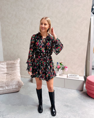 Model draagt een zwarte jurk met een bloemenpatroon. De jurk is getailleerd met een lintje en daaronder draagt ze lange zwarte botten.