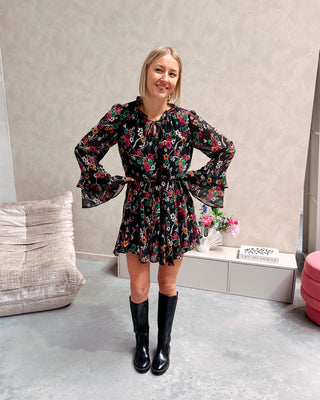 Model draagt een zwarte jurk met een bloemenpatroon. De jurk is getailleerd met een lintje en daaronder draagt ze lange zwarte botten.