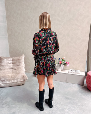 Model draagt een zwarte jurk met een bloemenpatroon. De jurk is getailleerd met een lintje en daaronder draagt ze lange zwarte botten.