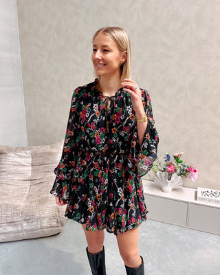 Model draagt een zwarte jurk met een bloemenpatroon. De jurk is getailleerd met een lintje en daaronder draagt ze lange zwarte botten.