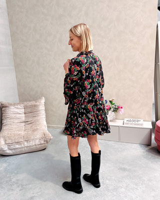 Model draagt een zwarte jurk met een bloemenpatroon. De jurk is getailleerd met een lintje en daaronder draagt ze lange zwarte botten.