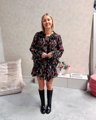 Model draagt een zwarte jurk met een bloemenpatroon. De jurk is getailleerd met een lintje en daaronder draagt ze lange zwarte botten.